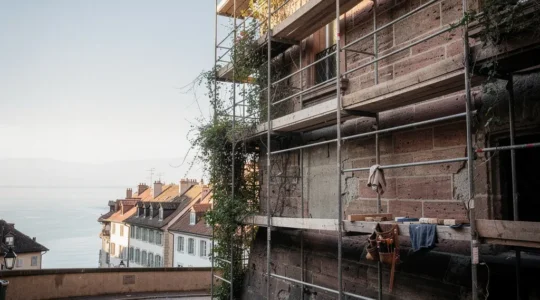 Façade en pierre de molasse d'un bâtiment historique de la Vieille-Ville de Genève avec des échafaudages de restauration visibles sous une lumière matinale douce